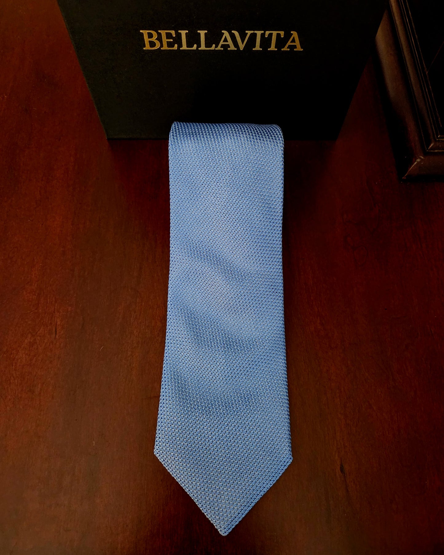 A light blue textured necktie laid flat on a dark brown wooden surface, with a black Bellavita box partially visible at the top.