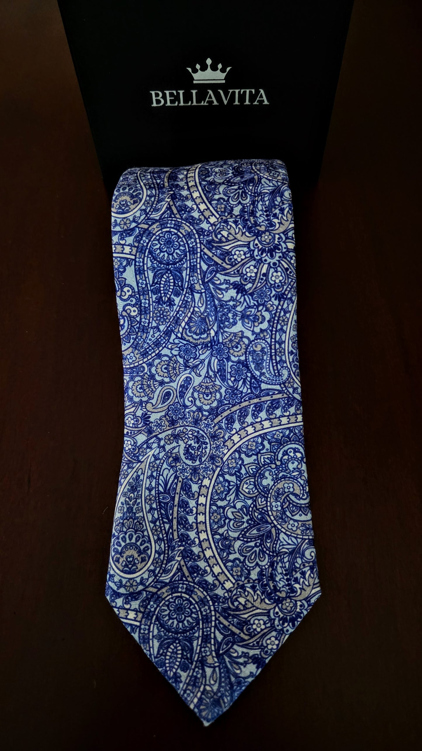 A blue and white paisley pattern necktie displayed vertically against a black Bellavita box on a dark brown background.
