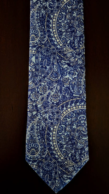 A classic necktie featuring an intricate blue and white paisley pattern, laid flat against a dark brown background.