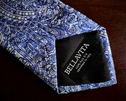 A close-up of a blue and white paisley patterned necktie with a black fabric brand label reading BELLAVITA.