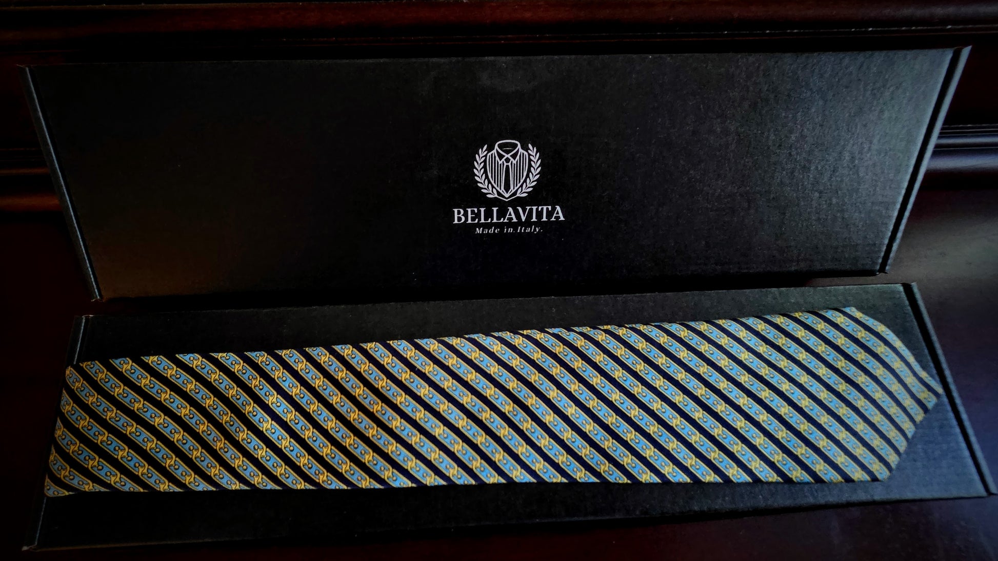 A Bellavita necktie with a blue, yellow, and gold geometric pattern, presented inside an open black gift box with the brand logo visible on the inner lid.