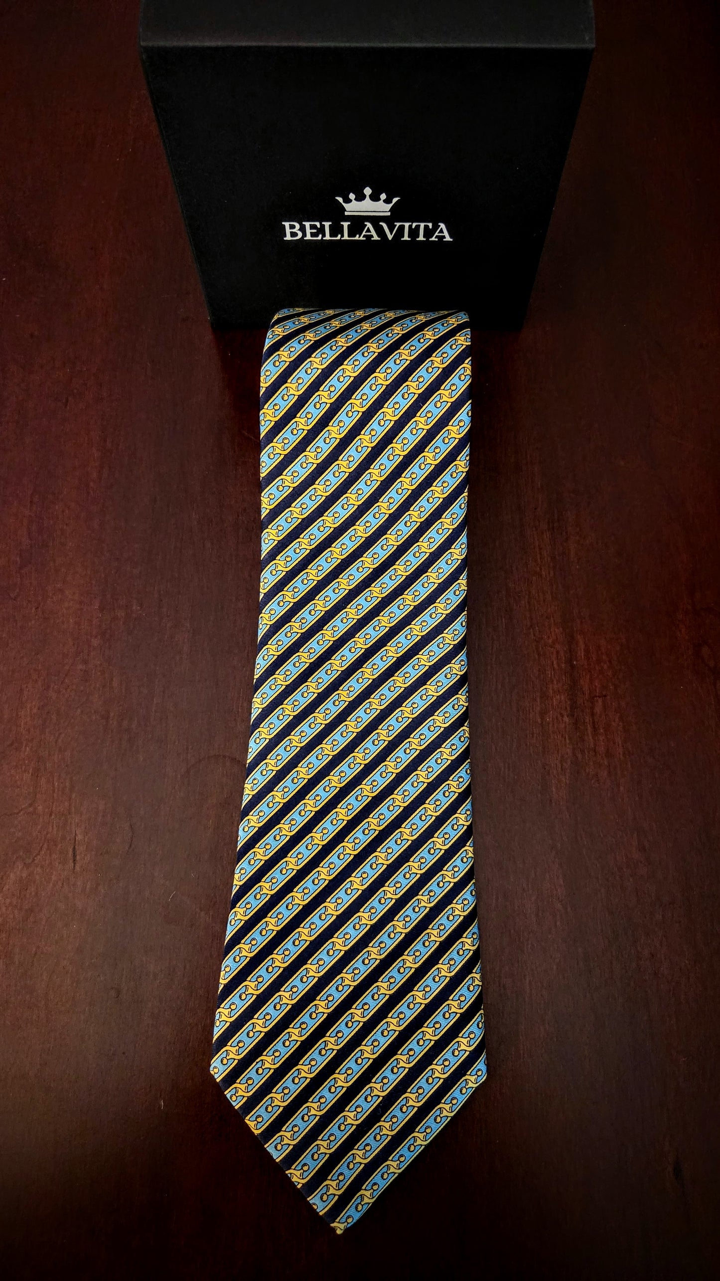 A Bellavita necktie with a diagonal striped pattern of yellow, blue, and white motifs on a black base, displayed vertically next to its black branded gift box on a dark wooden surface.