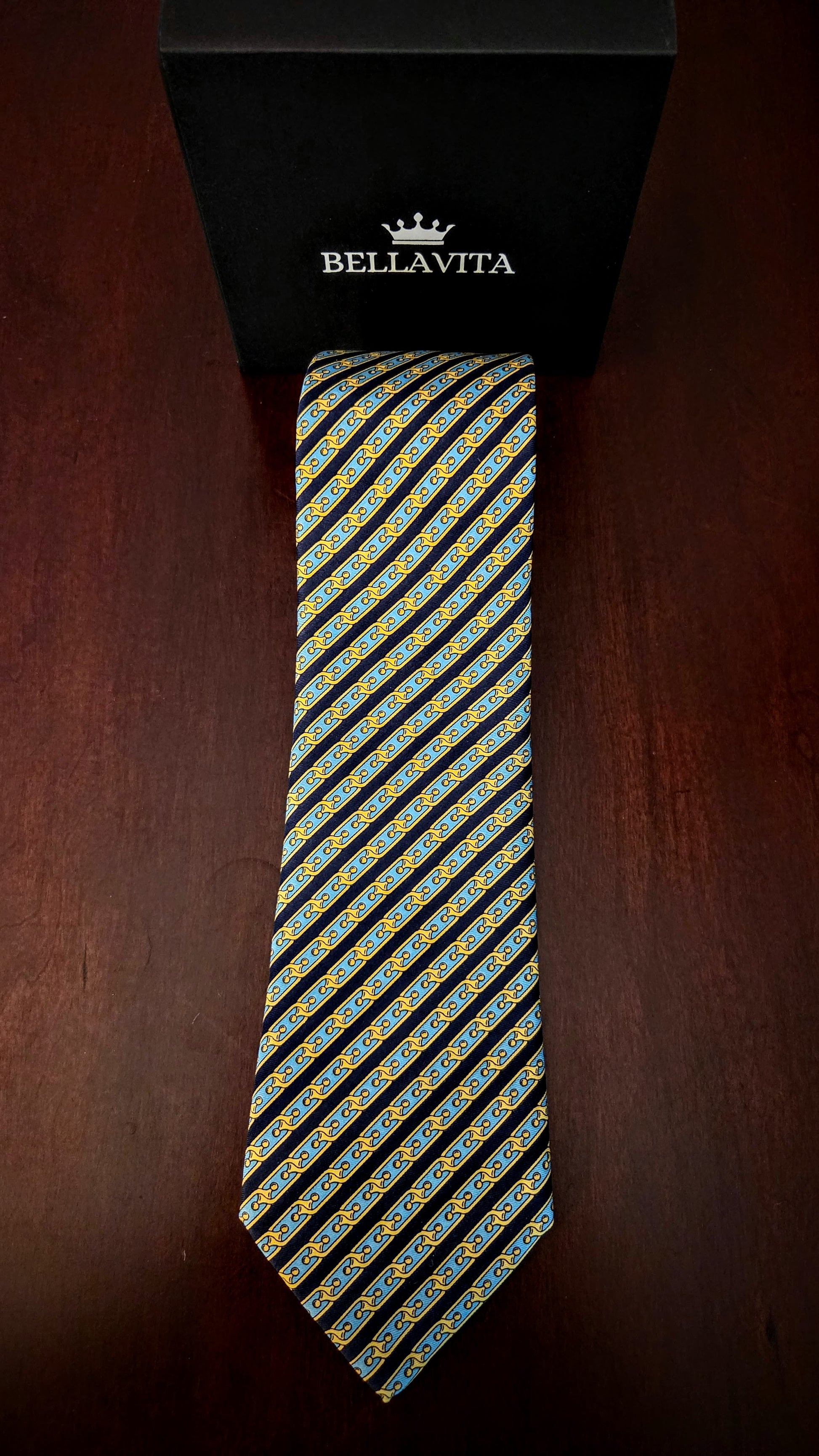 A Bellavita necktie with a diagonal striped pattern of yellow, blue, and white motifs on a black base, displayed vertically next to its black branded gift box on a dark wooden surface.