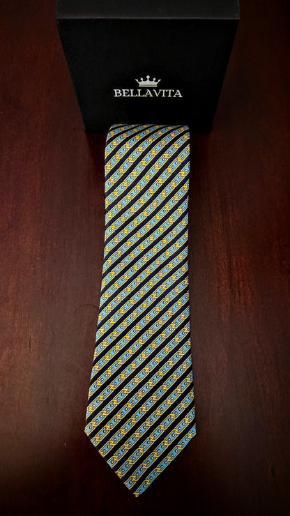 A Bellavita necktie with a diagonal striped pattern of yellow, blue, and white motifs on a black base, displayed vertically next to its black branded gift box on a dark wooden surface.