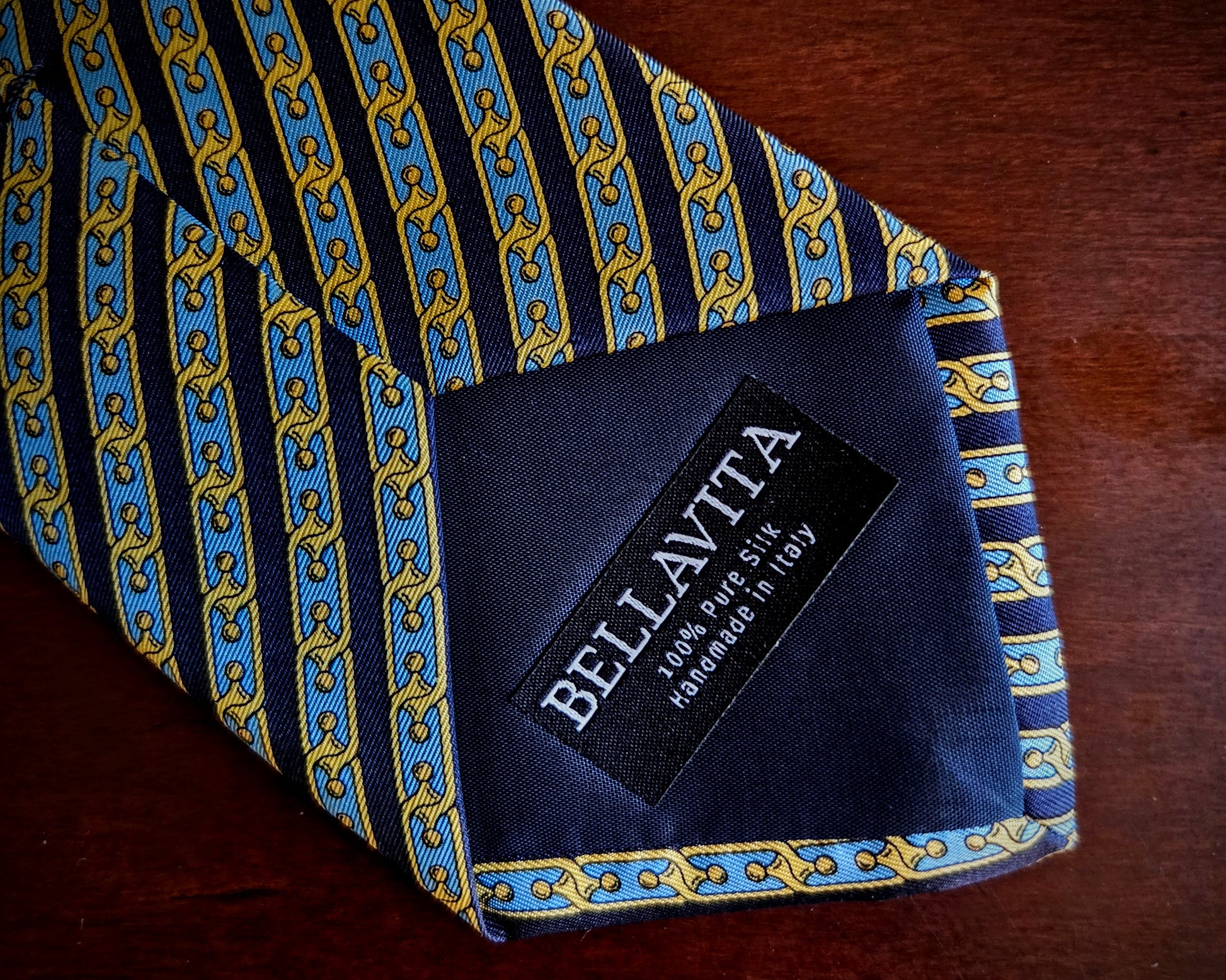 A close-up of a Bellavita necktie on a dark wooden surface, showing a blue and gold chain-link pattern with a black brand label.