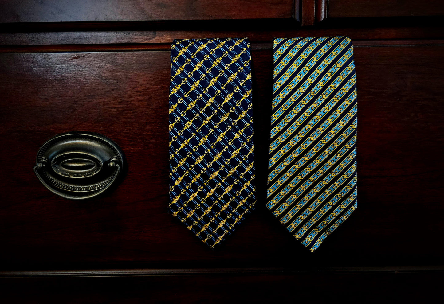 Two patterned neckties hanging on a dark wooden drawer front, with a metal drawer pull visible on the left. One tie has a navy and gold geometric pattern, and the other has a blue, yellow, and green striped pattern.