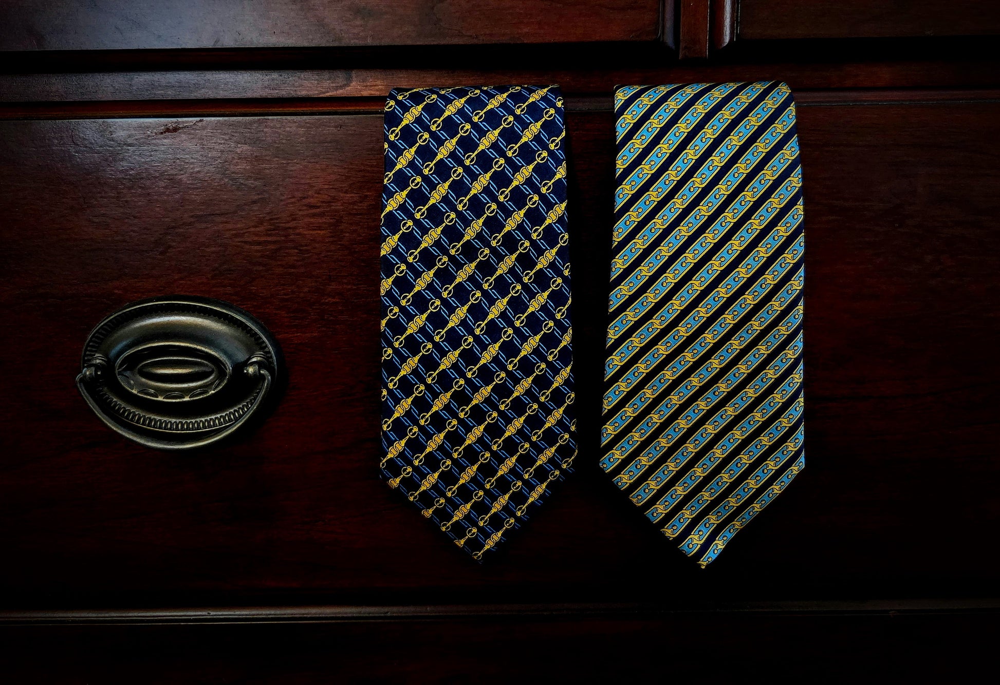 Two patterned neckties hanging on a dark wooden drawer front, with a metal drawer pull visible on the left. One tie has a navy and gold geometric pattern, and the other has a blue, yellow, and green striped pattern.