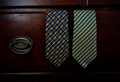 Two patterned neckties hanging on a dark wooden drawer front, with a metal drawer pull visible on the left. One tie has a navy and gold geometric pattern, and the other has a blue, yellow, and green striped pattern.