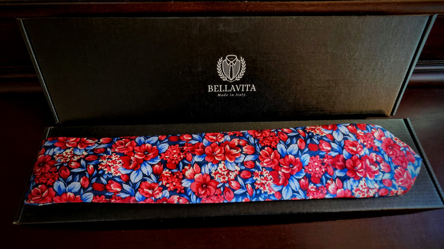 A Bellavita necktie with a vibrant red and blue floral pattern, displayed inside its black presentation box on a dark wooden surface.