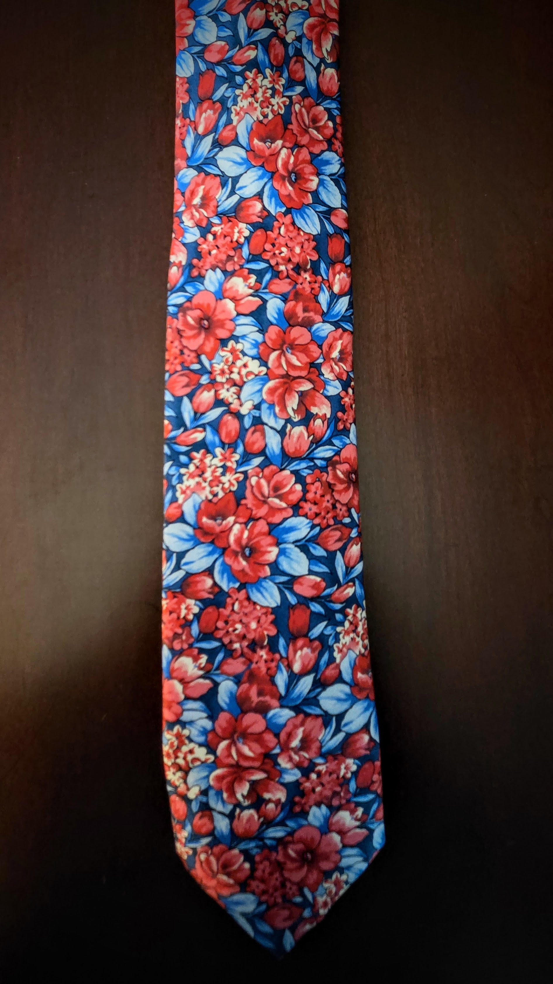 A necktie with a vibrant red and blue floral pattern, laid flat against a dark brown background.