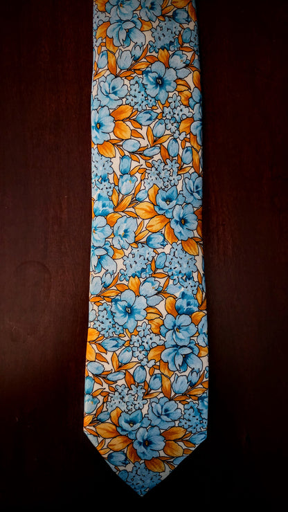 A necktie with a dense floral pattern in light blue, orange, and yellow on a white base, displayed vertically against a dark brown background.
