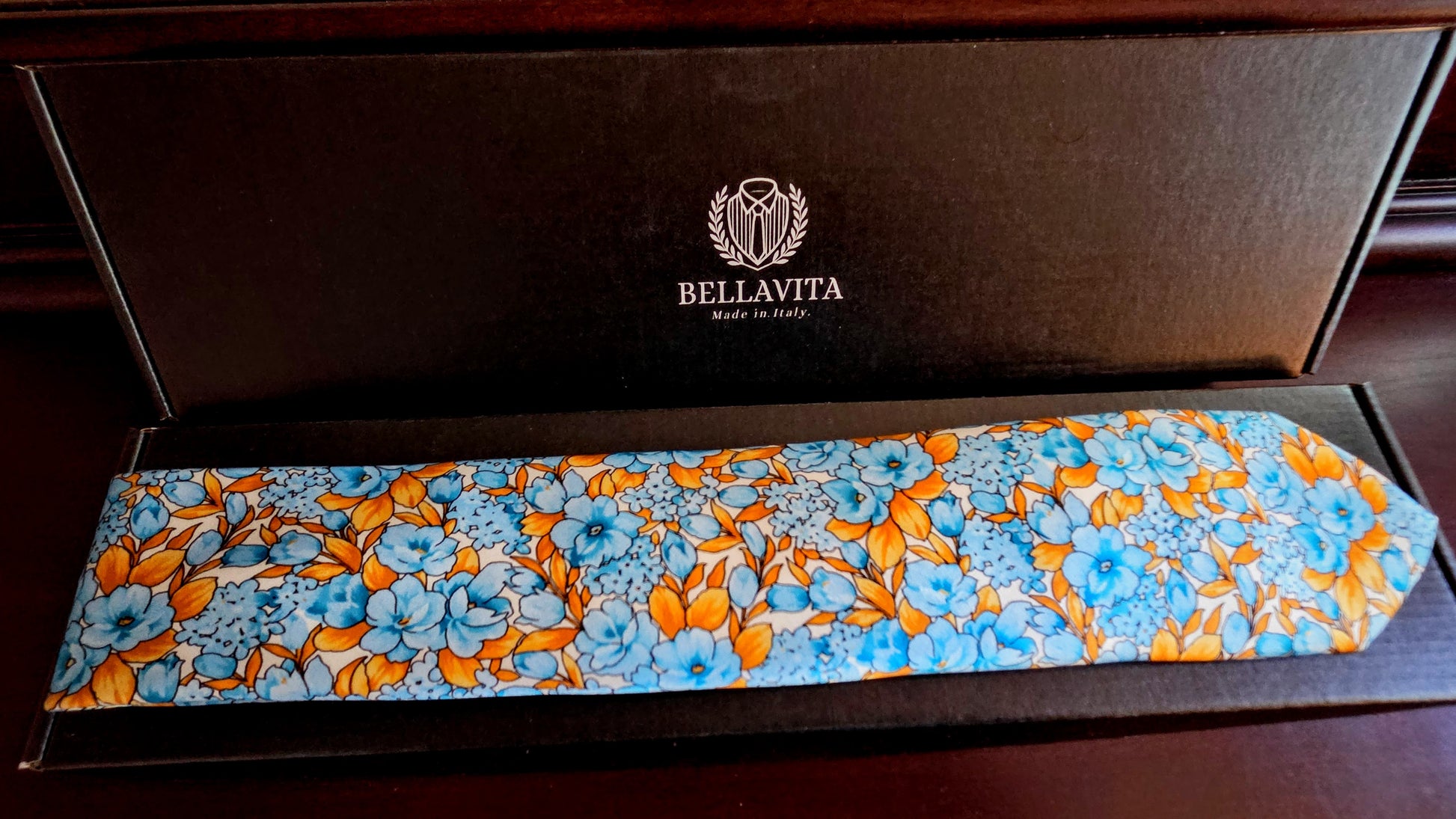 A Bellavita silk necktie with a vibrant blue and orange floral pattern, presented inside its dark brown branded gift box.