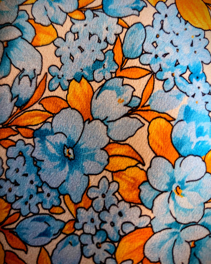 A close-up of a vintage-style floral fabric or wallpaper pattern featuring light blue and orange flowers with black outlines on a cream background.