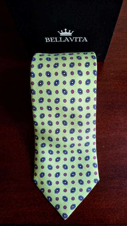 A light green silk necktie with a repeating blue and red circular pattern, displayed on a dark wooden surface in front of a black Bellavita box.