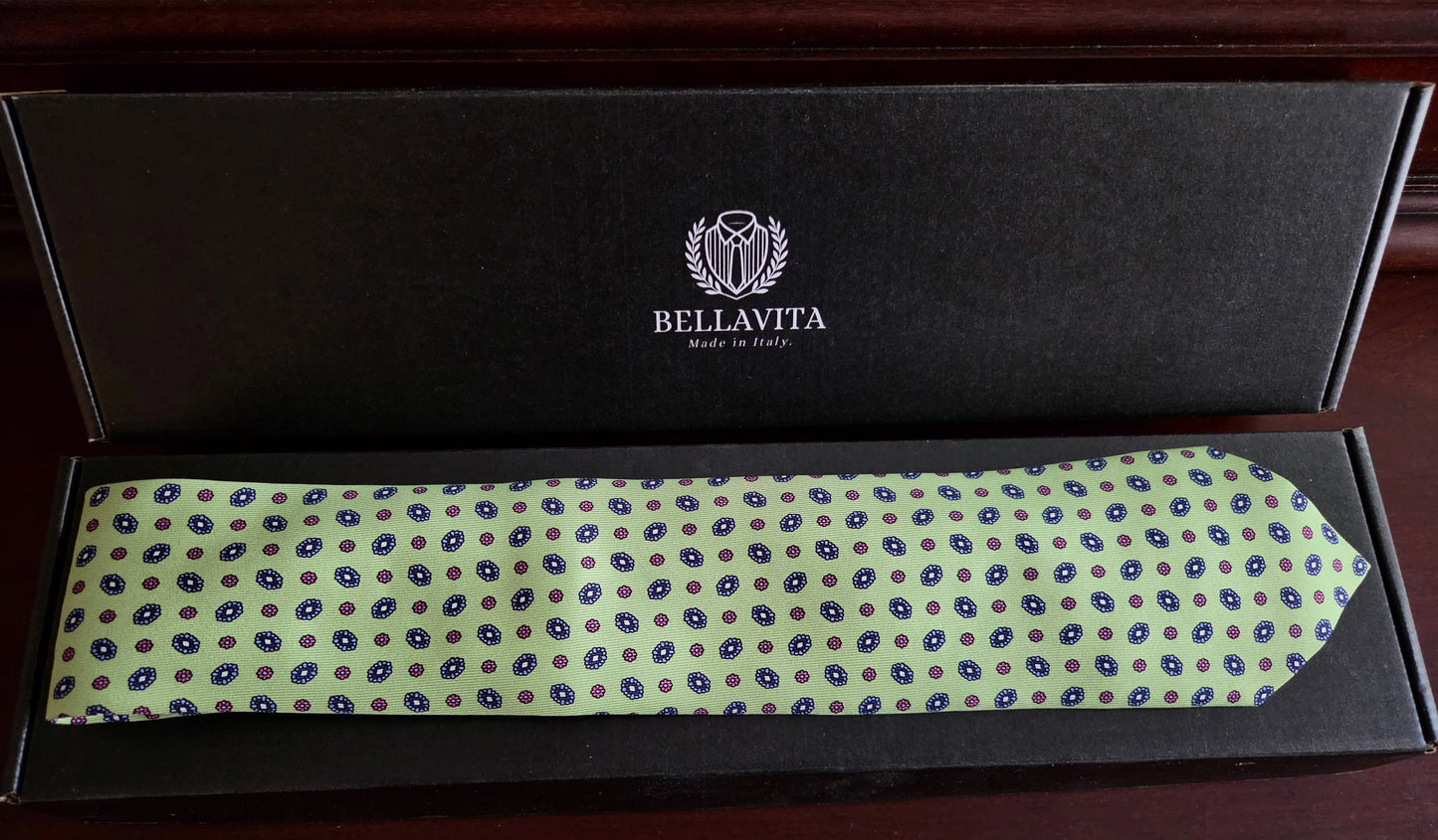 A Bellavita necktie with a light green base and blue and brown polka dots, presented inside its black branded box on a dark wooden surface.