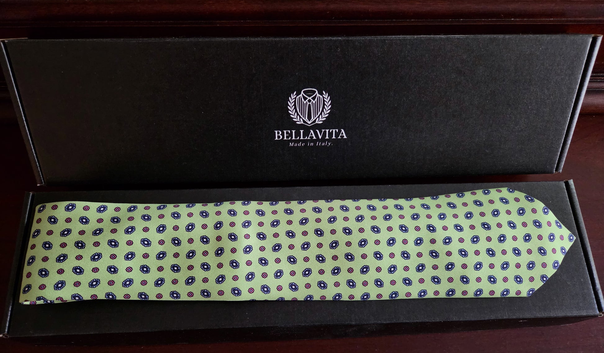 A Bellavita necktie with a light green base and blue and brown polka dots, presented inside its black branded box on a dark wooden surface.