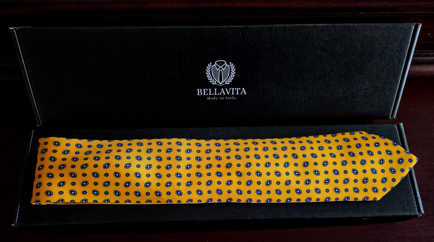 A yellow necktie with a blue polka-dot pattern, presented inside an open black Bellavita gift box on a dark wooden surface.