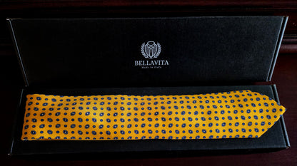 A yellow necktie with a blue polka-dot pattern, presented inside an open black Bellavita gift box on a dark wooden surface.