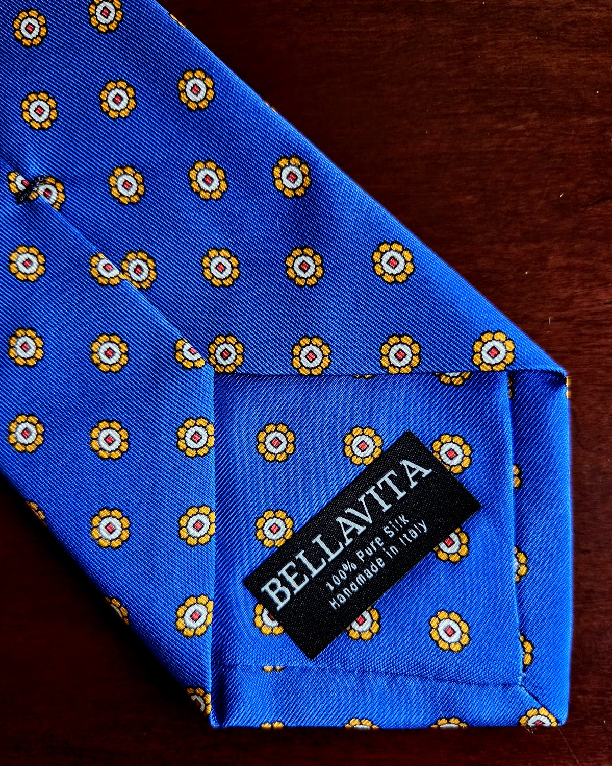 A close-up of a blue silk necktie with a repeating yellow and white floral pattern, featuring a black fabric brand label that reads 'BELLAVITA'.
