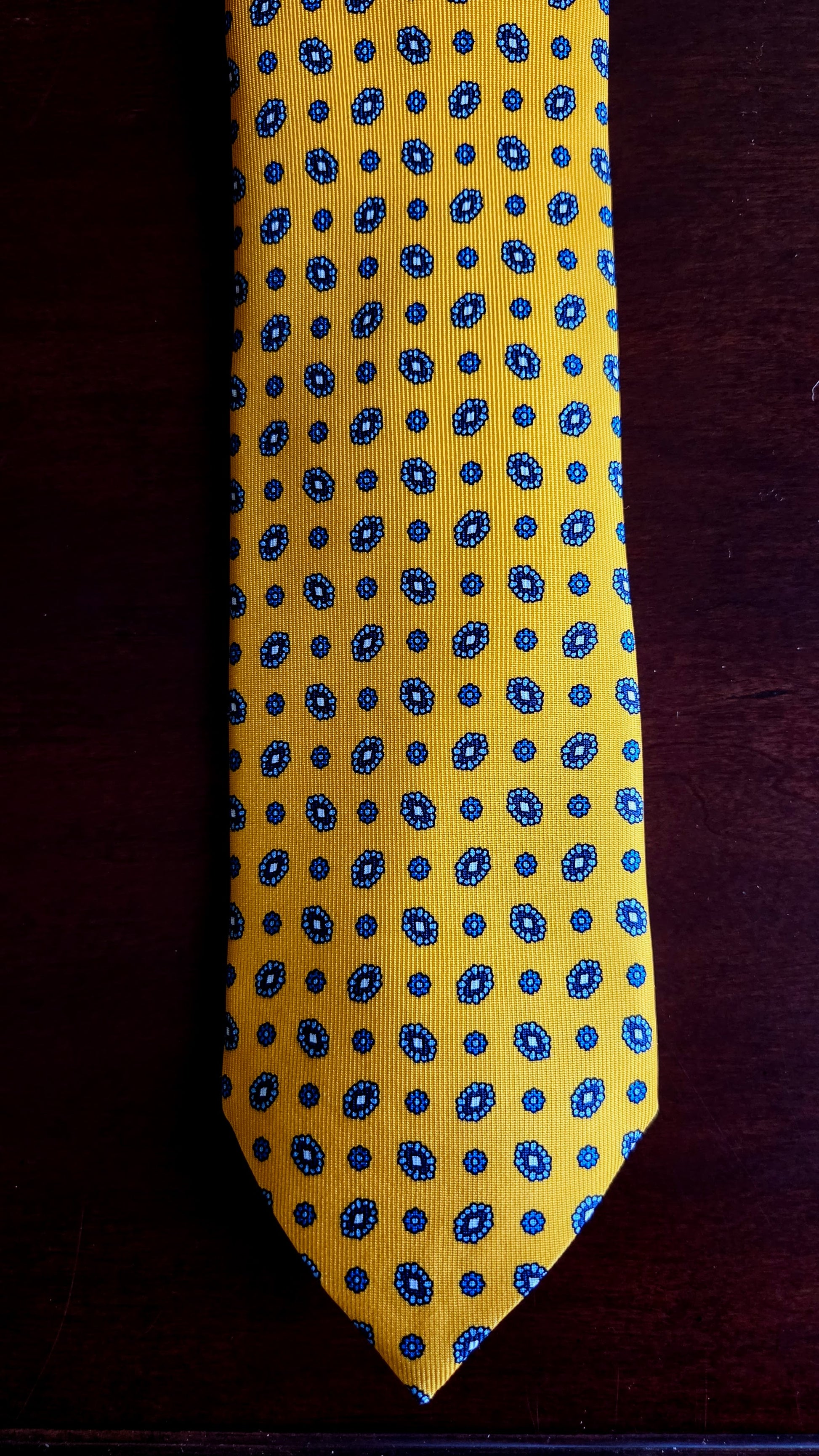 A yellow necktie with a repeating blue and white paisley pattern, laid flat on a dark brown wooden surface.