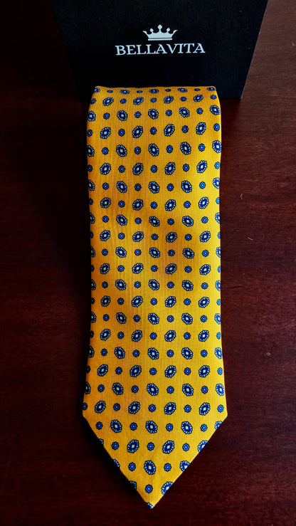 A yellow silk necktie with a blue and white geometric pattern, displayed vertically next to its black Bellavita brand box on a dark wooden surface.