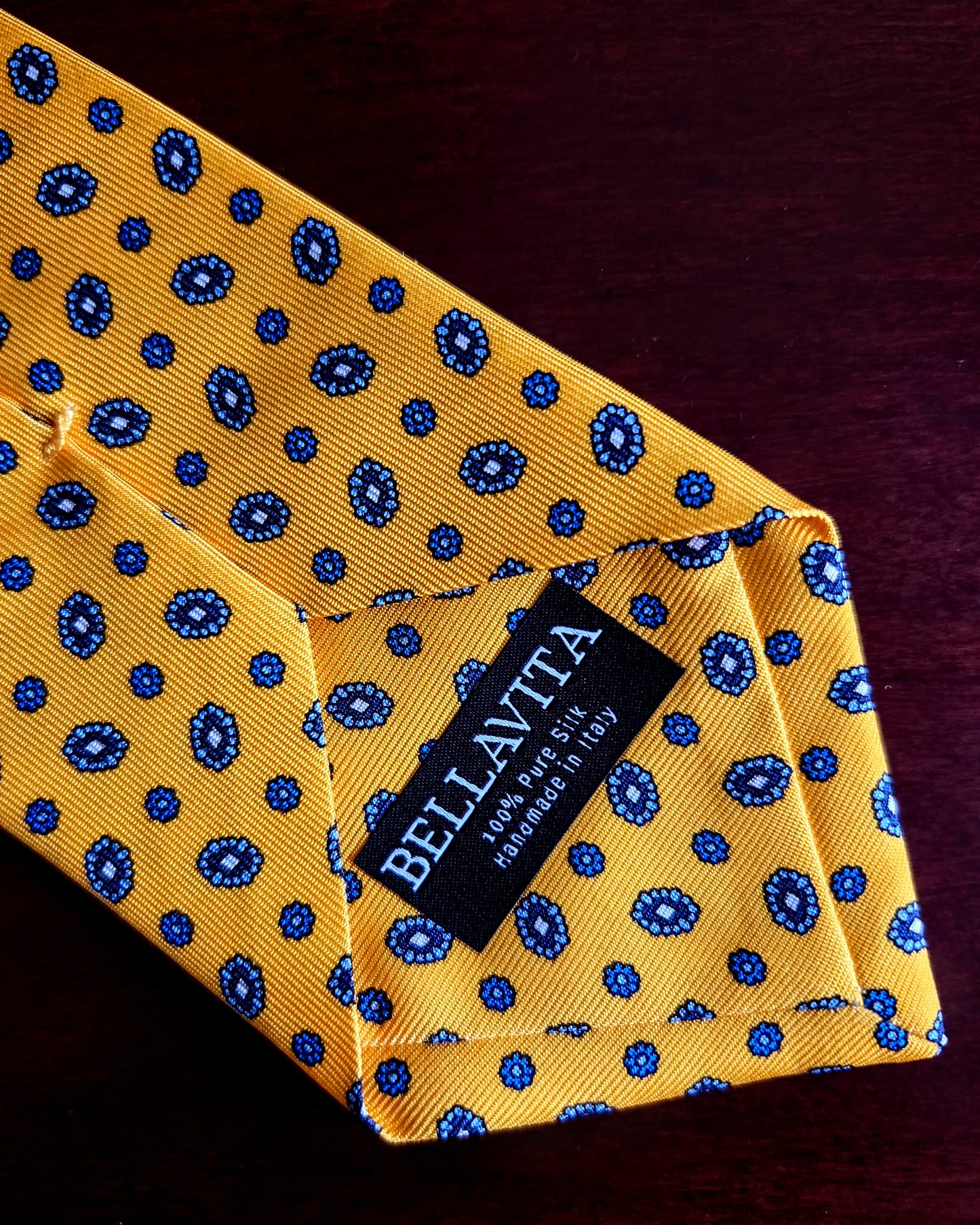 A close-up of a yellow silk necktie with a blue paisley pattern, folded to show a black fabric brand label that reads 'BELLAVITA'.