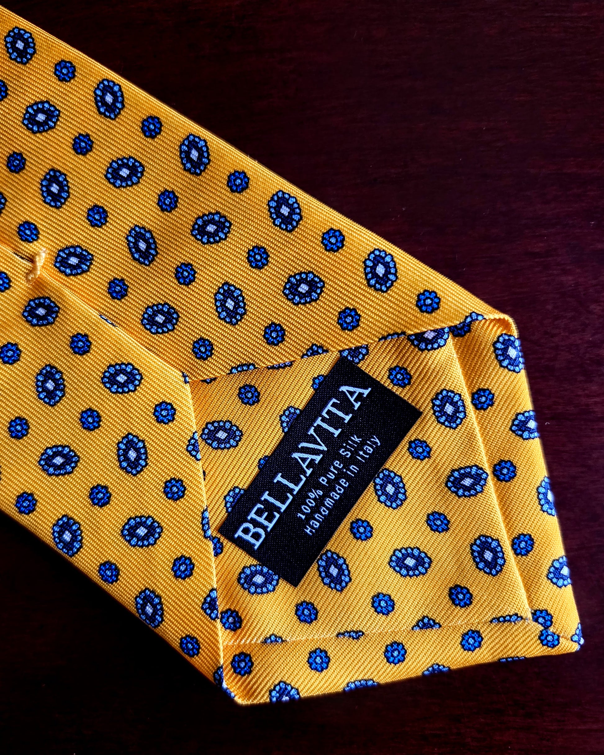 A close-up of a yellow silk necktie with a blue paisley pattern, folded to show a black fabric brand label that reads 'BELLAVITA'.