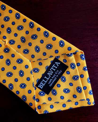 A close-up of a yellow silk necktie with a blue paisley pattern, folded to show a black fabric brand label that reads 'BELLAVITA'.