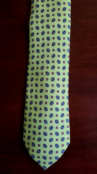A light green necktie with a repeating pattern of blue and red circular medallions, laid flat on a dark brown wooden surface.