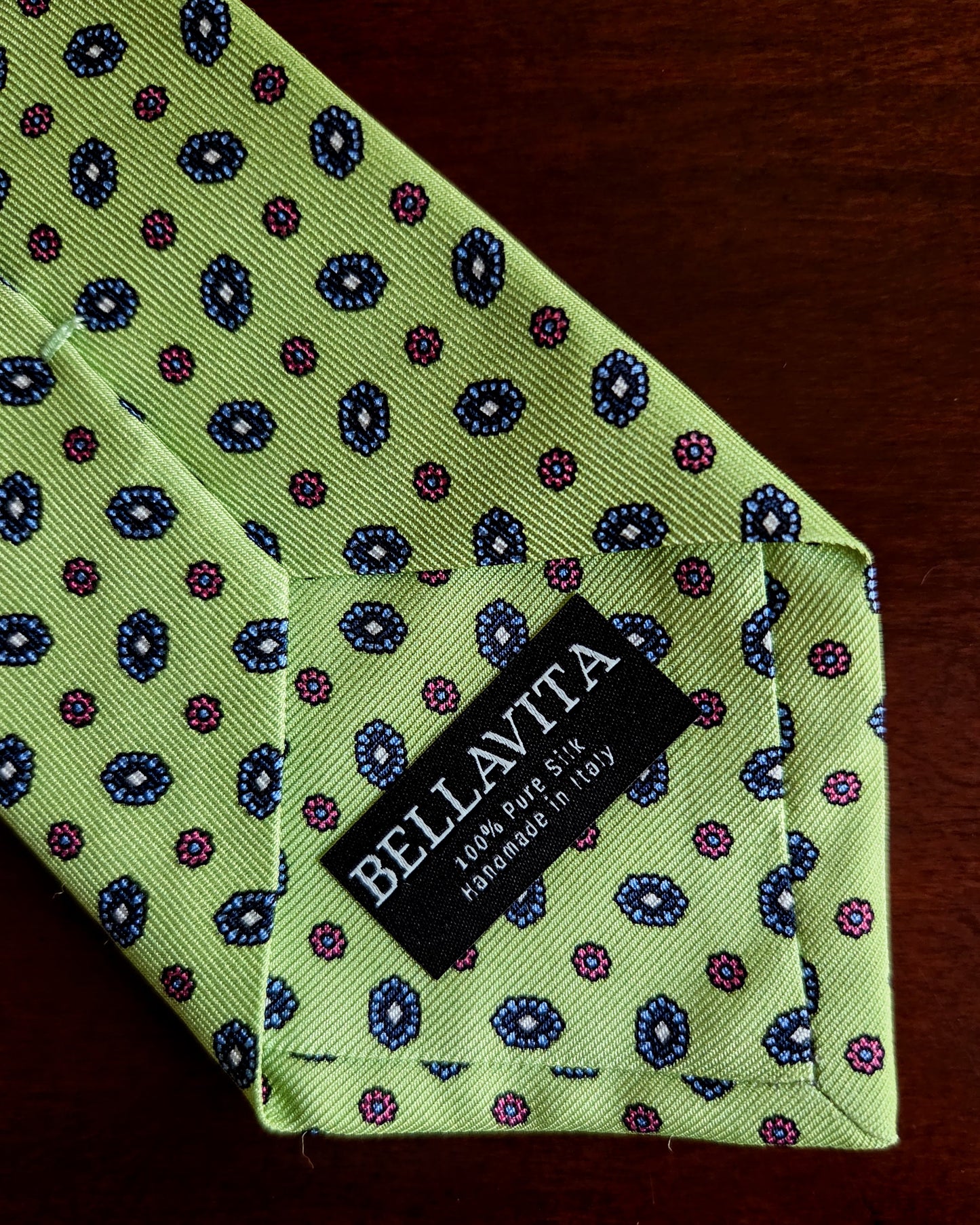 A close-up of a Bellavita necktie in light green silk with a repeating paisley pattern in navy blue and magenta, laid flat on a dark wooden surface.