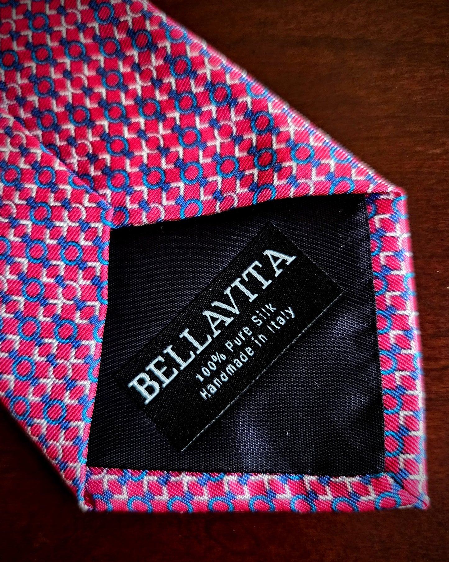 A close-up of a Bellavita silk necktie with a pink, blue, and white geometric pattern, showing the black interior label with white text.
