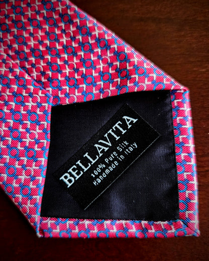 A close-up of a Bellavita silk necktie with a pink, blue, and white geometric pattern, showing the black interior label with white text.