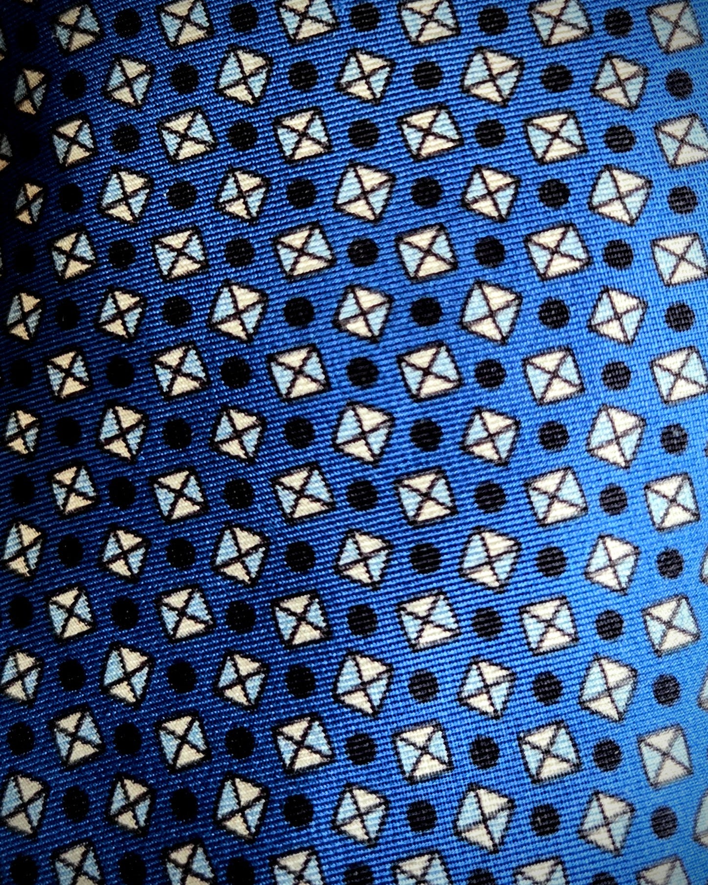 A close-up of a patterned fabric, likely a necktie, featuring a repeating design of white and light blue geometric shapes on a bright blue background with small black dots.