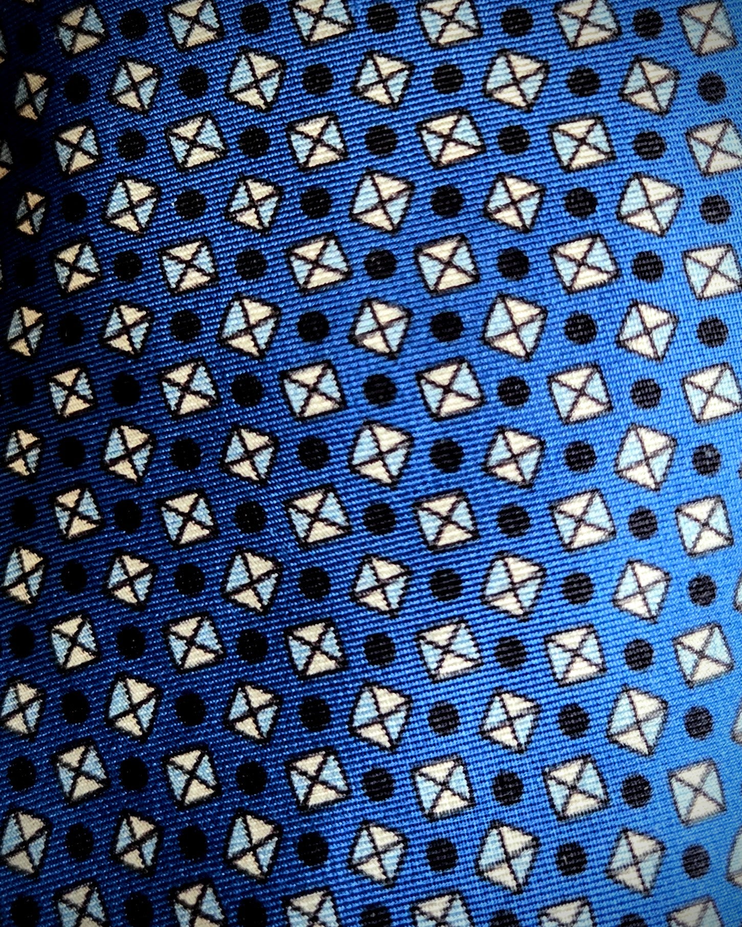 A close-up of a patterned fabric, likely a necktie, featuring a repeating design of white and light blue geometric shapes on a bright blue background with small black dots.