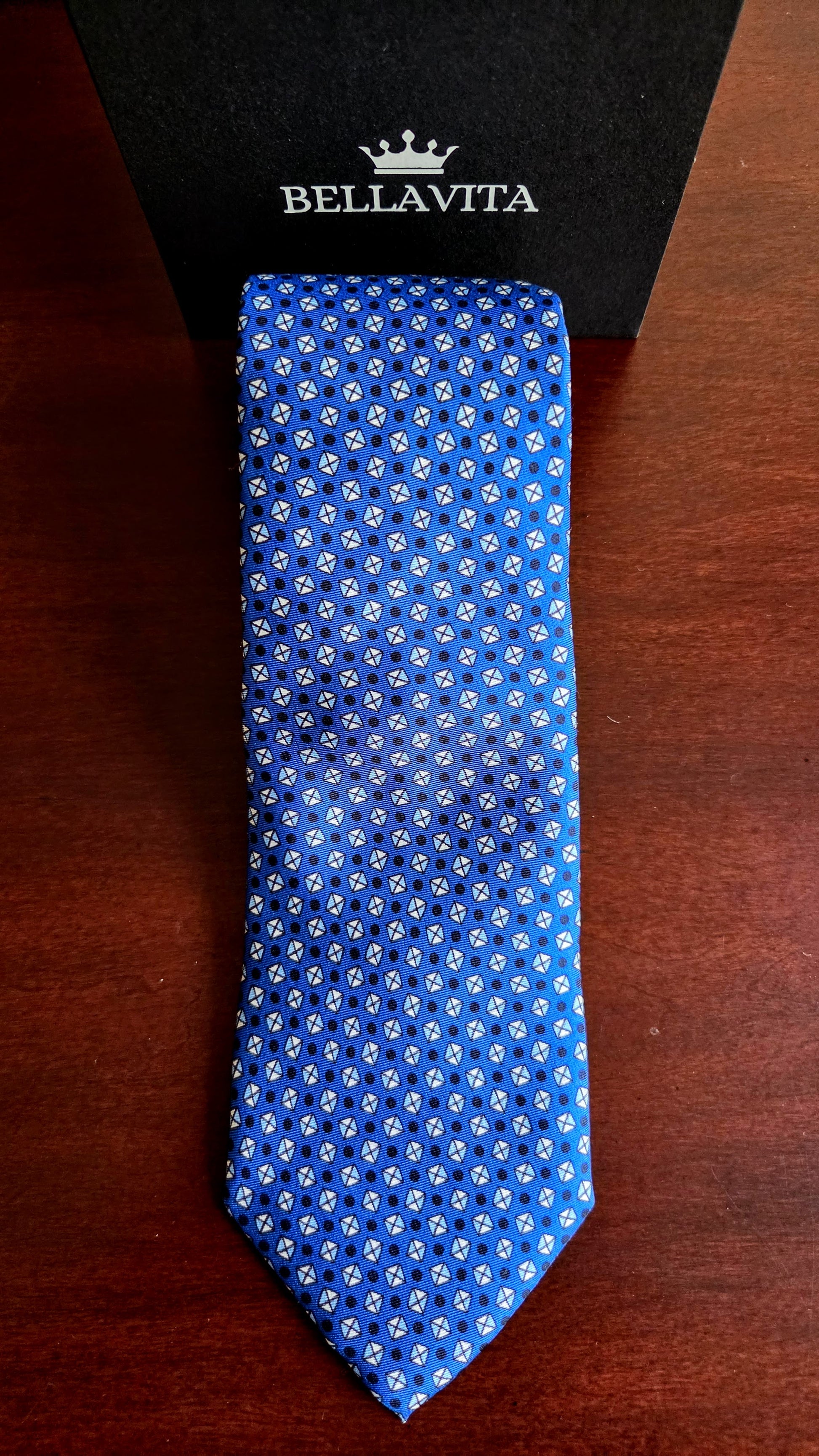 A blue silk necktie with a white geometric pattern, displayed vertically next to its black Bellavita brand box on a dark wooden surface.