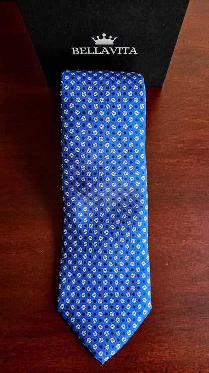 A blue silk necktie with a white geometric pattern, displayed vertically next to its black Bellavita brand box on a dark wooden surface.
