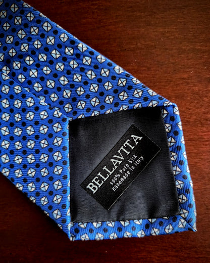 A close-up of a blue necktie with a repeating white and black geometric pattern, laid on a dark wooden surface. The black fabric neckband shows the brand name 'BELLAVITA' in white text.