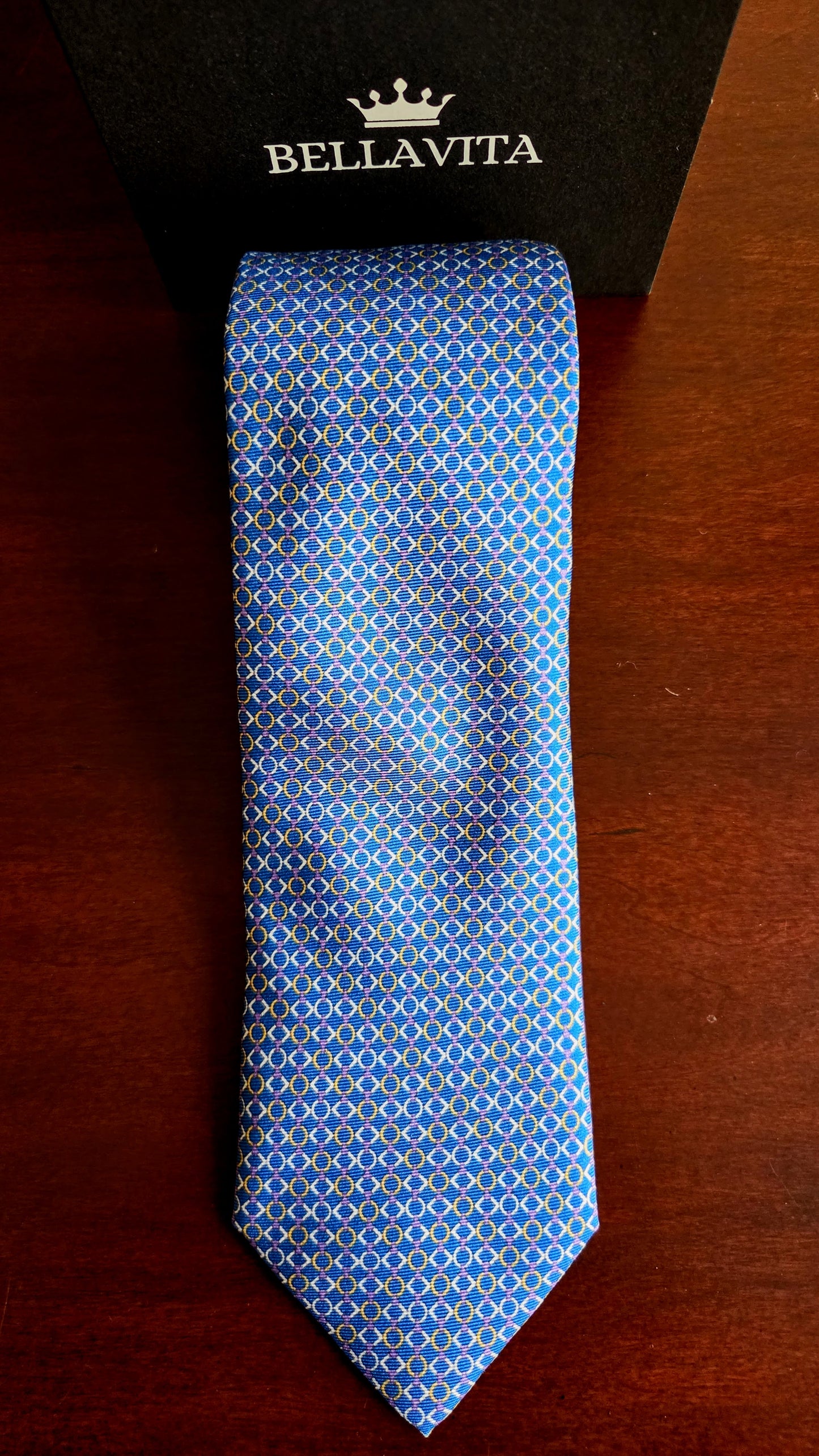A blue patterned silk necktie with a repeating geometric design, laid flat on a dark wooden surface in front of its black Bellavita box.