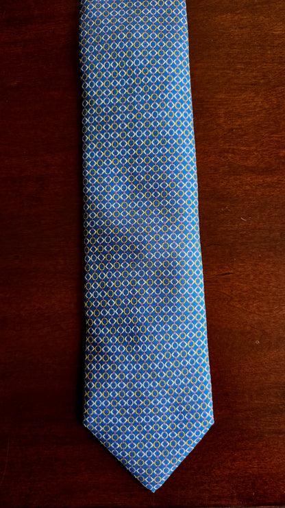 A blue necktie with a repeating geometric diamond pattern in yellow and white, laid flat on a dark brown wooden surface.