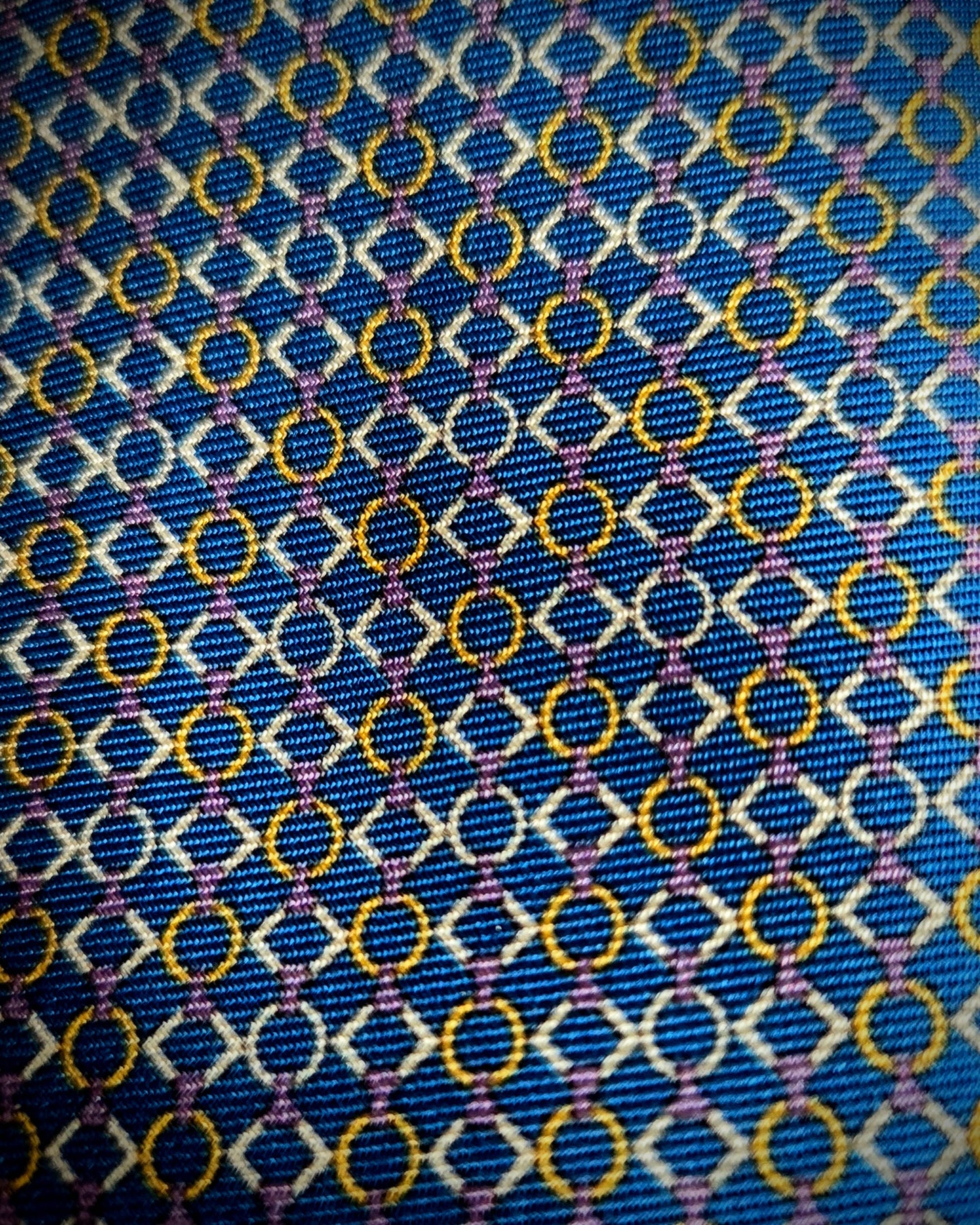 A close-up of a patterned fabric with a blue base, featuring repeating white diamond outlines, yellow-gold circular motifs, and thin purple intersecting lines.