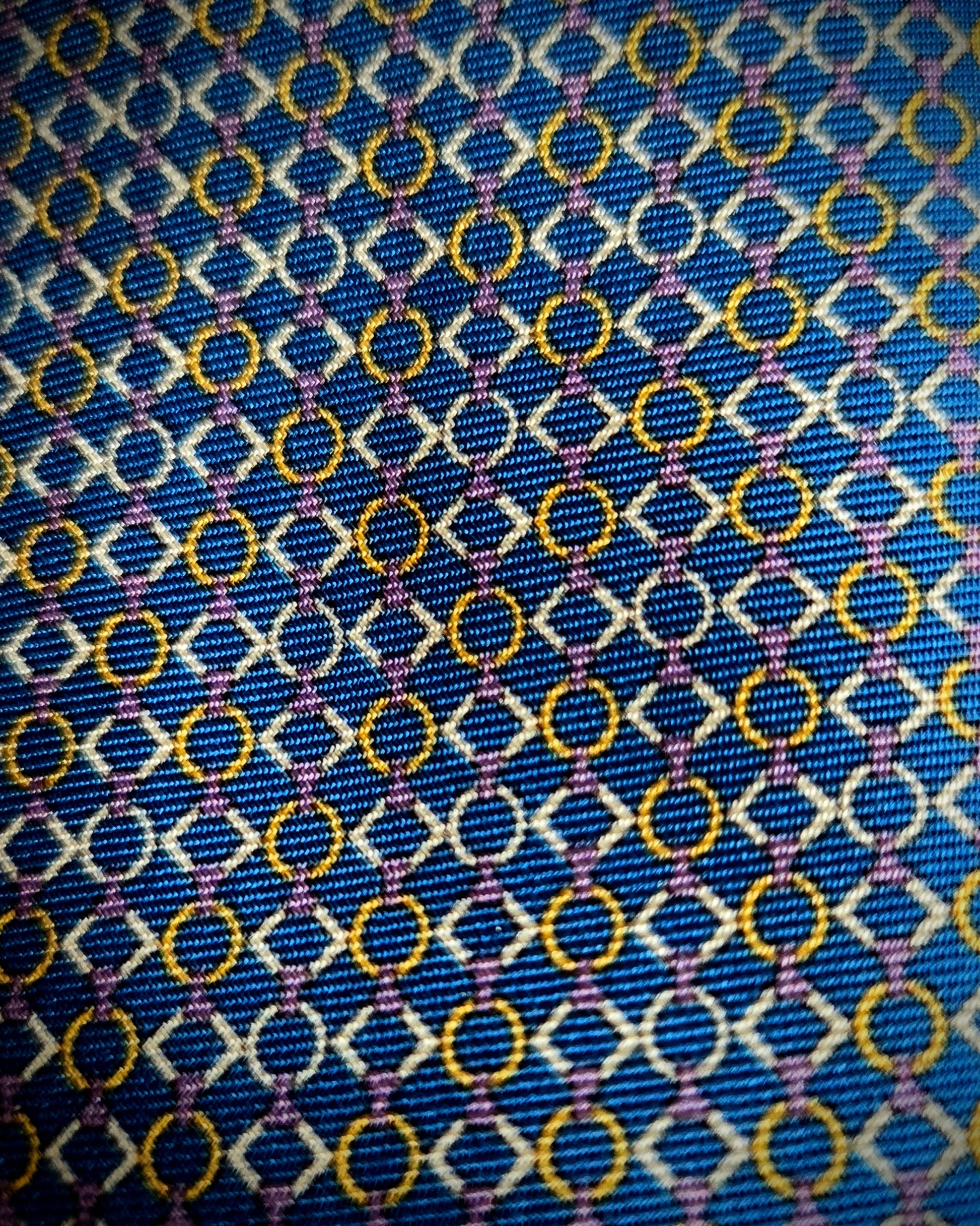 A close-up of a patterned fabric with a blue base, featuring repeating white diamond outlines, yellow-gold circular motifs, and thin purple intersecting lines.