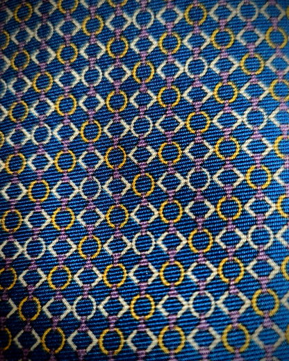 A close-up of a patterned fabric with a blue base, featuring repeating white diamond outlines, yellow-gold circular motifs, and thin purple intersecting lines.