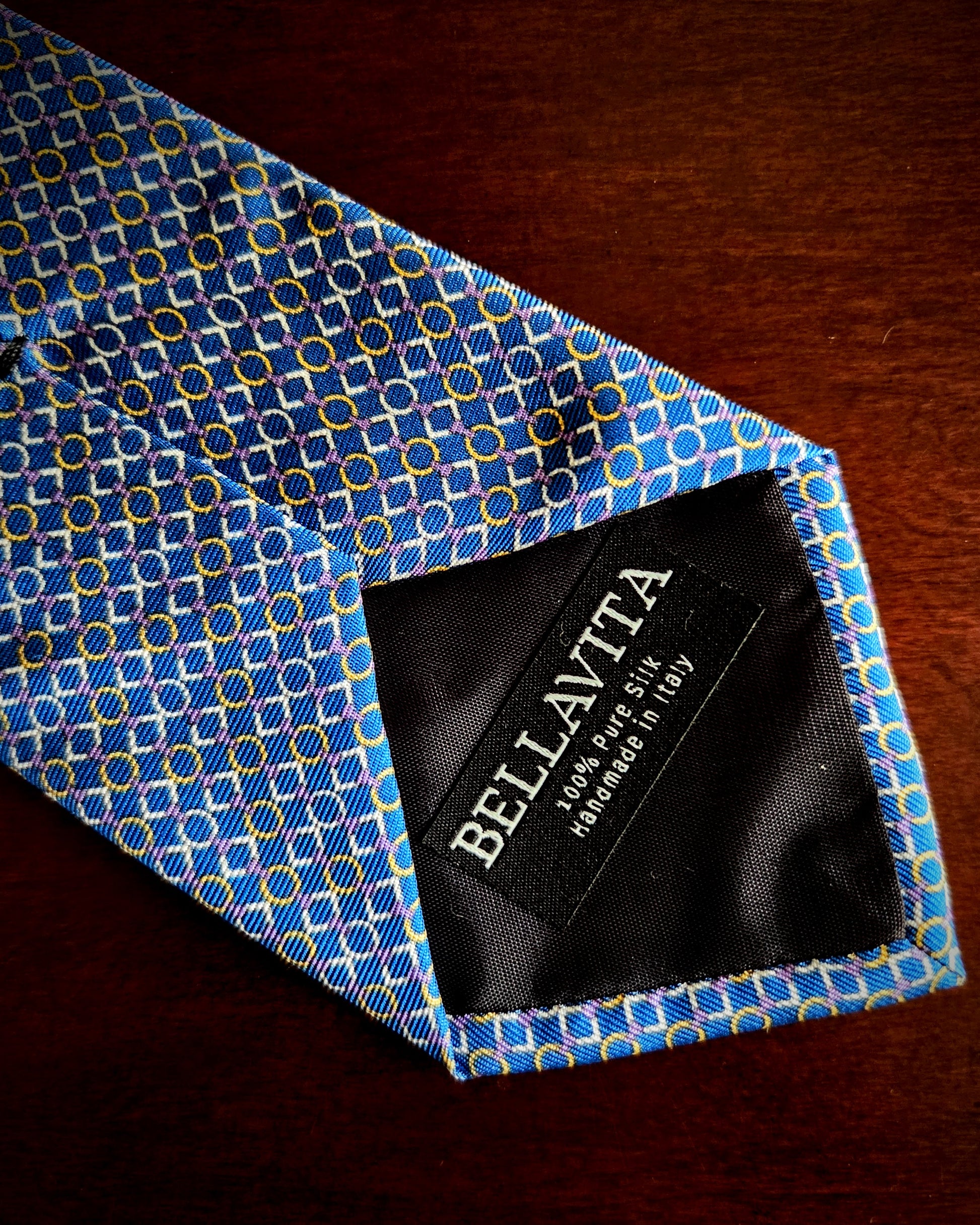 A close-up of a blue patterned necktie with yellow and white geometric details, laid on a dark wooden surface. The inside neckband is visible with a black label reading BELLAVITA.