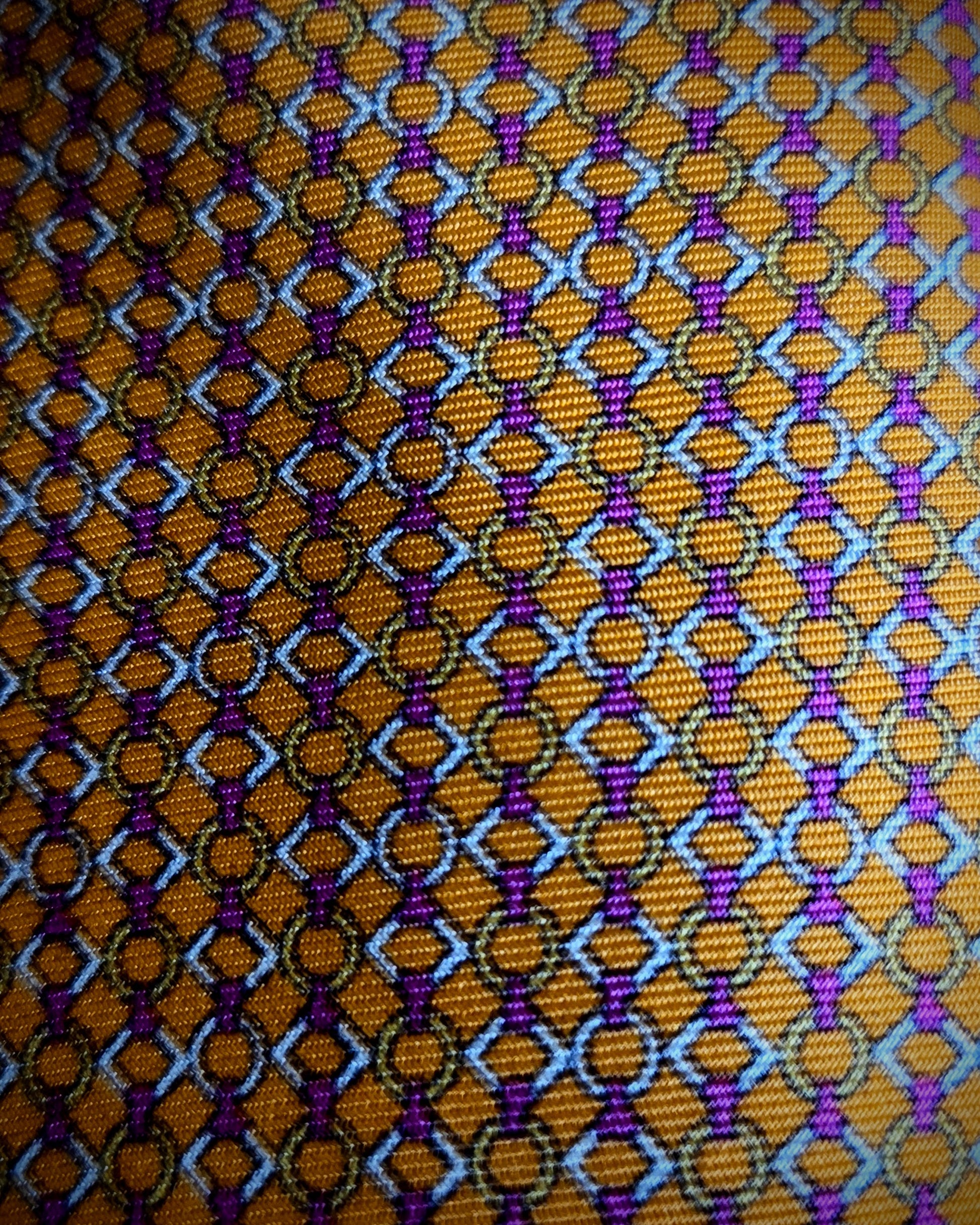 A close-up of a woven fabric pattern featuring a repeating geometric design in orange, purple, light blue, and black.