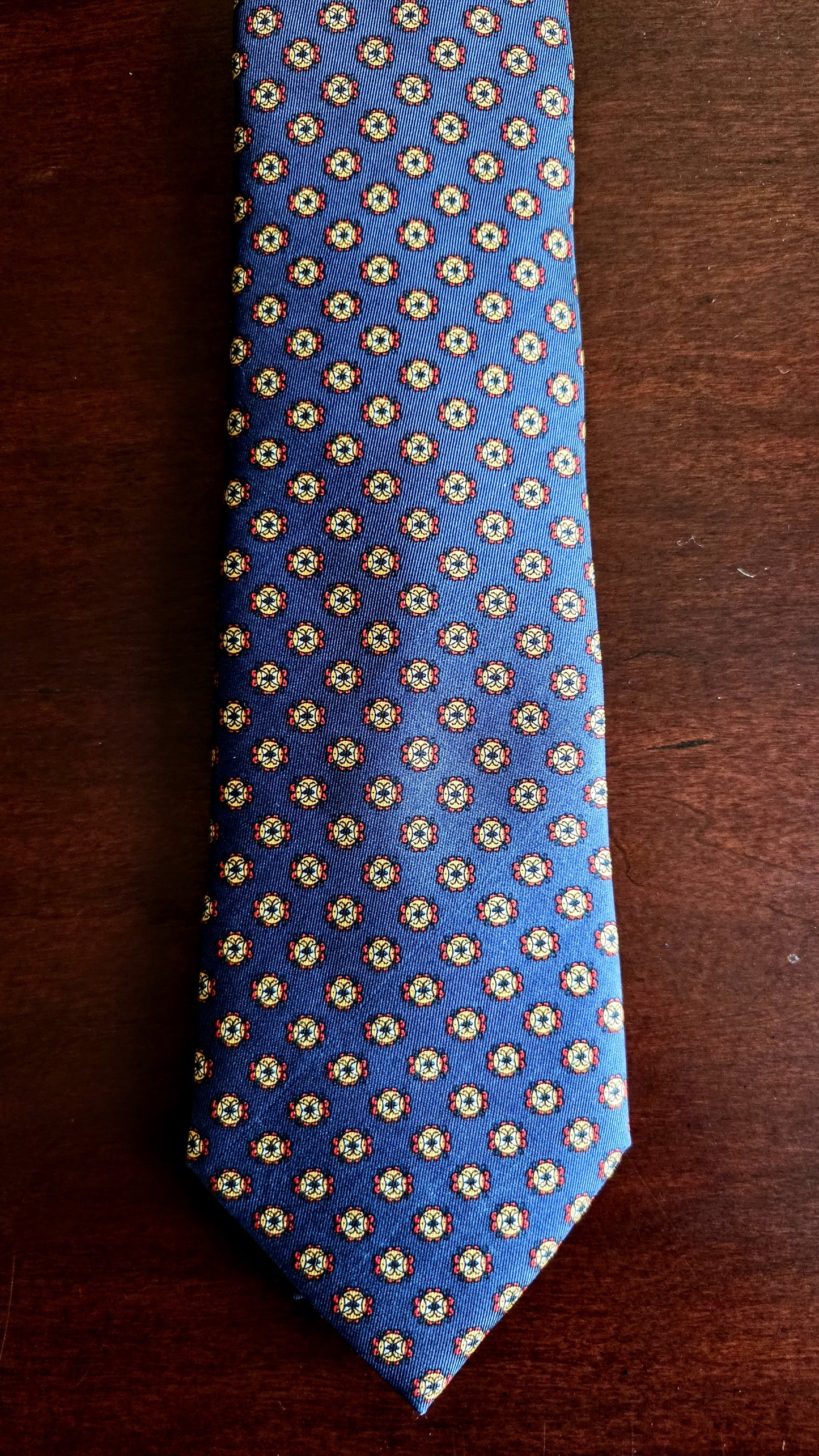 A blue necktie with a repeating small geometric pattern in beige and white, laid flat on a dark brown wooden surface.