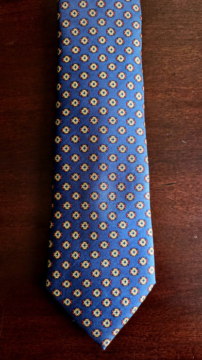 A blue necktie with a repeating small geometric pattern in beige and white, laid flat on a dark brown wooden surface.