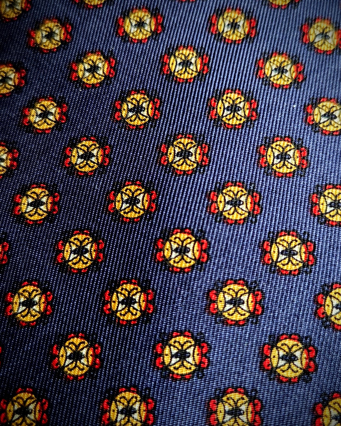 A close-up of a woven fabric pattern featuring a repeating medallion motif in yellow, red, black, and white on a dark navy blue background.