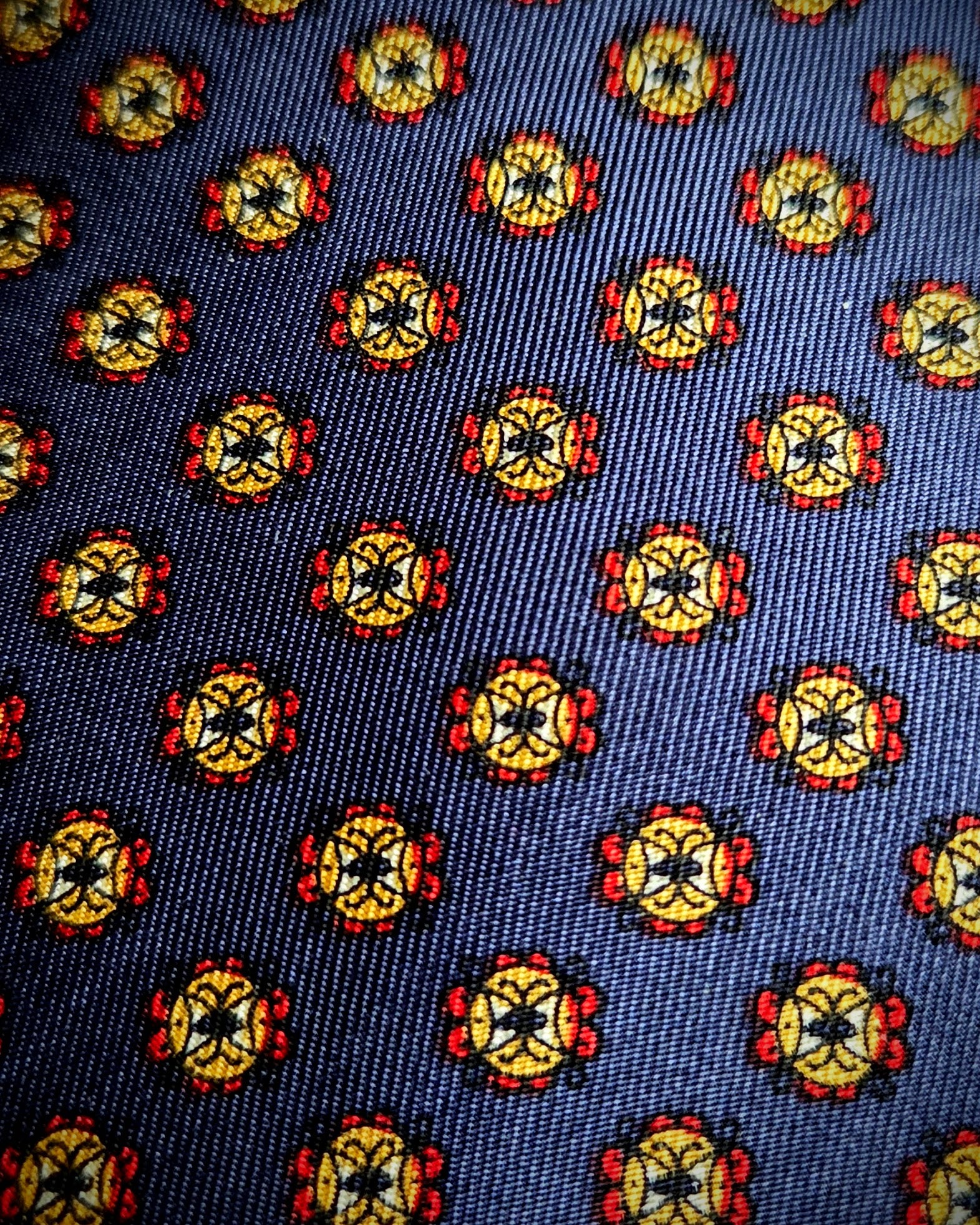 A close-up of a woven fabric pattern featuring a repeating medallion motif in yellow, red, black, and white on a dark navy blue background.
