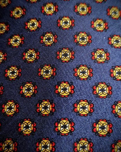 A close-up of a woven fabric pattern featuring a repeating medallion motif in yellow, red, black, and white on a dark navy blue background.
