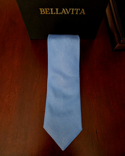 A light blue textured necktie laid flat on a dark brown wooden surface, with a black Bellavita box partially visible at the top.